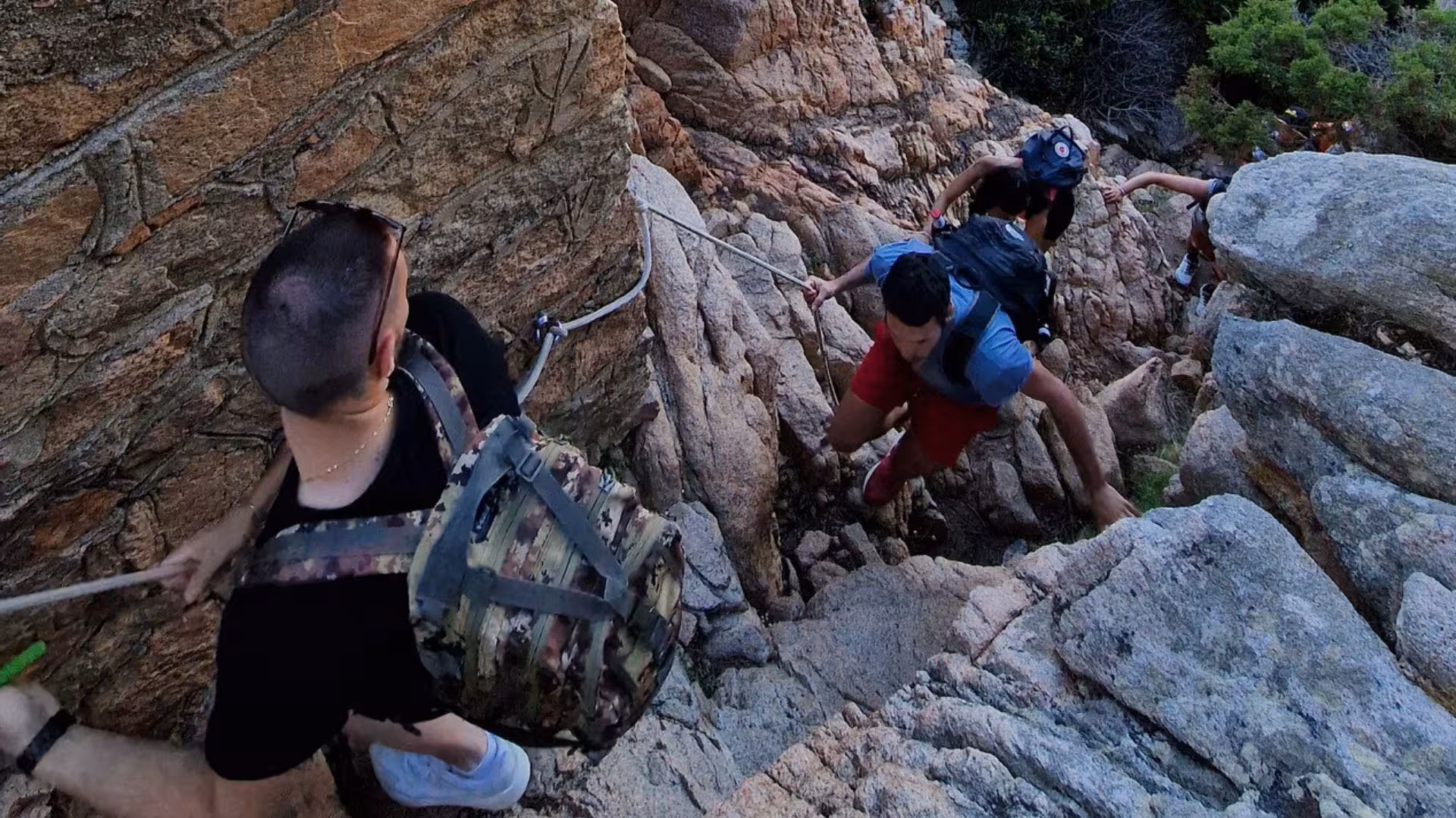 Adventurous hikers navigating steep rocky path with support ropes on Caprera Island.