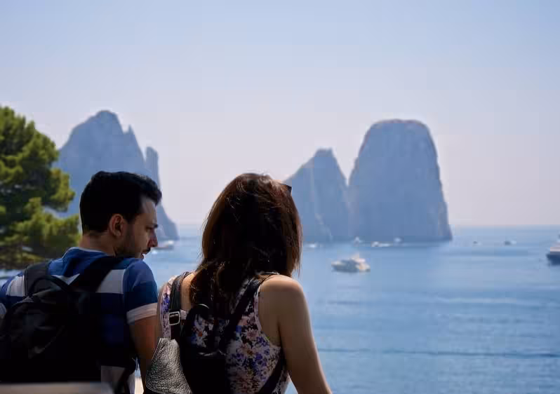 Couple enjoying a panoramic view of Capri's Faraglioni rock formations and serene sea under a clear sky.
