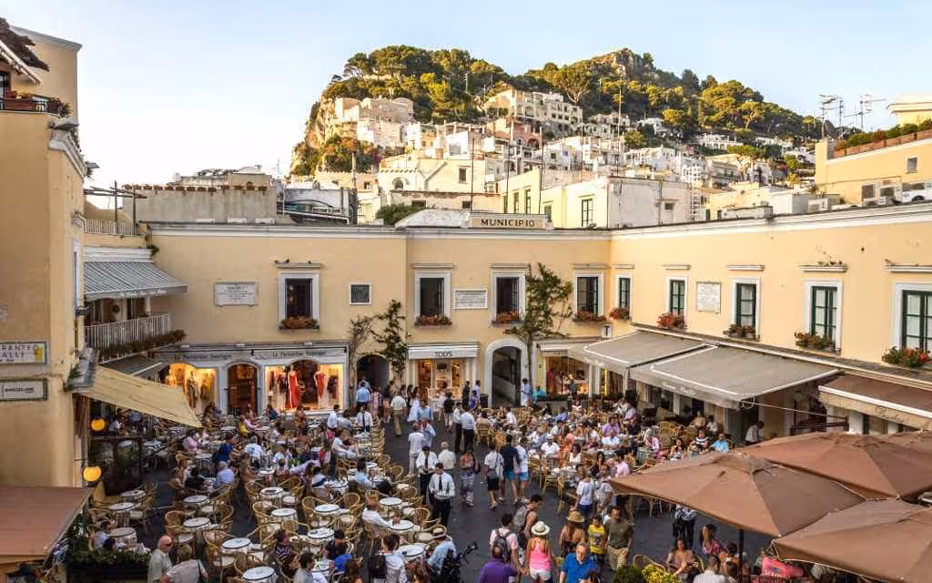 Bustling Capri square with outdoor cafes surrounded by charming architecture and lush hillside backdrop.