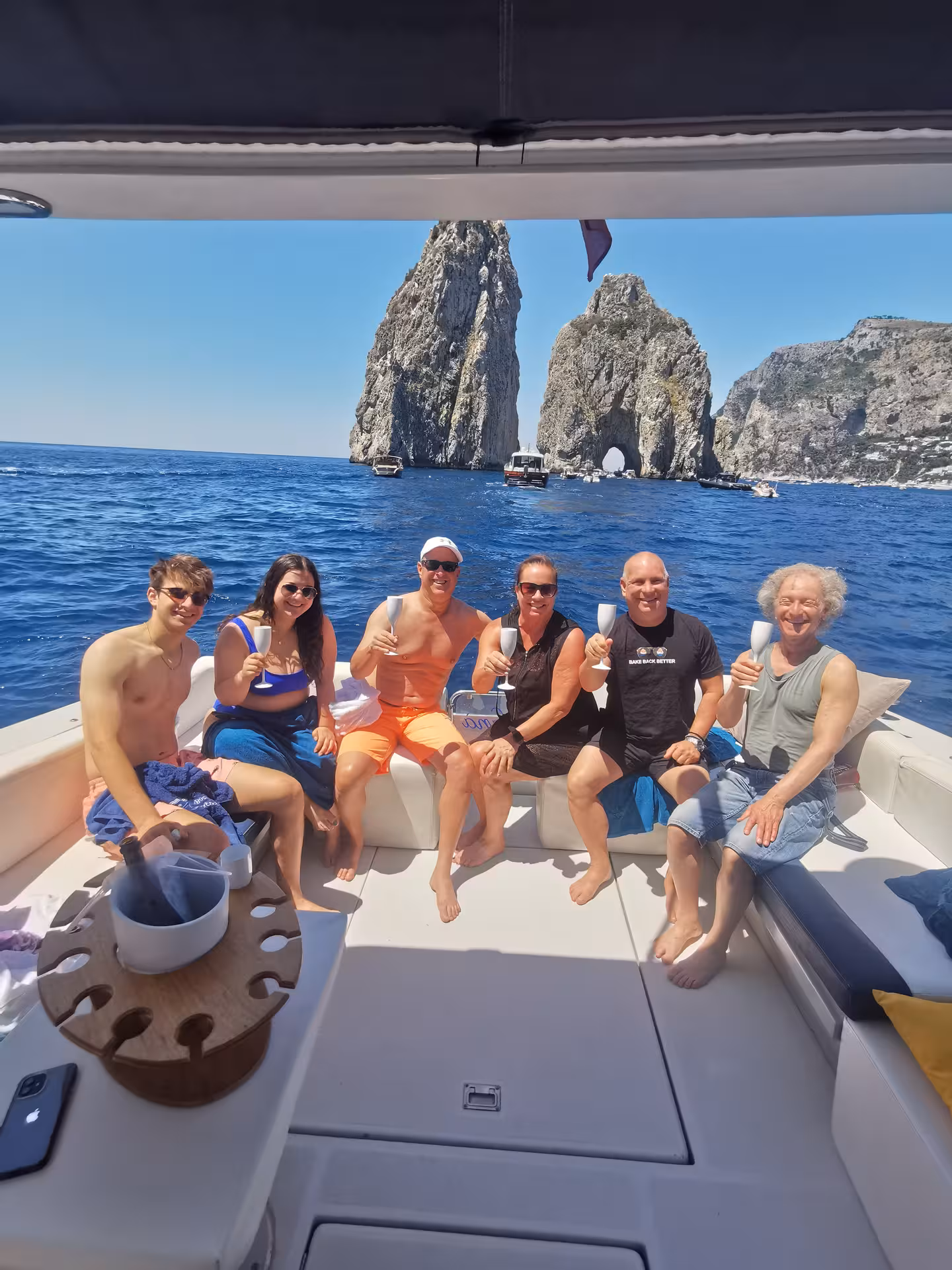 Group of friends enjoying limoncello on a boat with Capri's iconic Faraglioni rocks in the background.