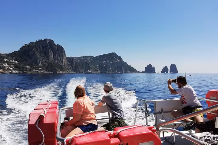 Tourists enjoy scenic views of Capri's Faraglioni rocks from a boat, capturing the picturesque seascape on camera.