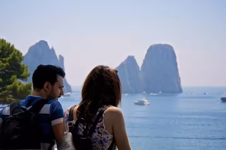Tourists enjoying a scenic view of Capri's Faraglioni rocks from a vantage point on a sunny day.