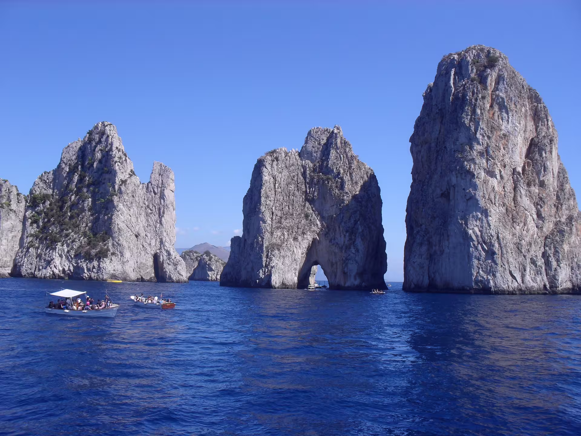 Small tour boats cruising around Capri’s iconic Faraglioni sea stacks on a sunny day trip in southern Italy