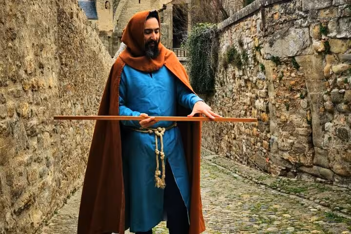 Medieval guide in blue tunic and brown hooded cloak holding a wooden measuring stick in Carcassonne stone alley