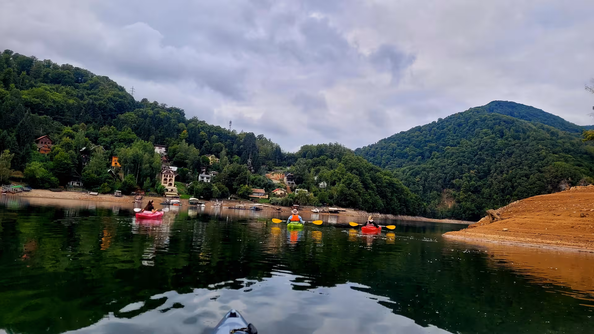 Kayakers paddling on a mountain lake surrounded by forested Carpathian hills and a small lakeside village