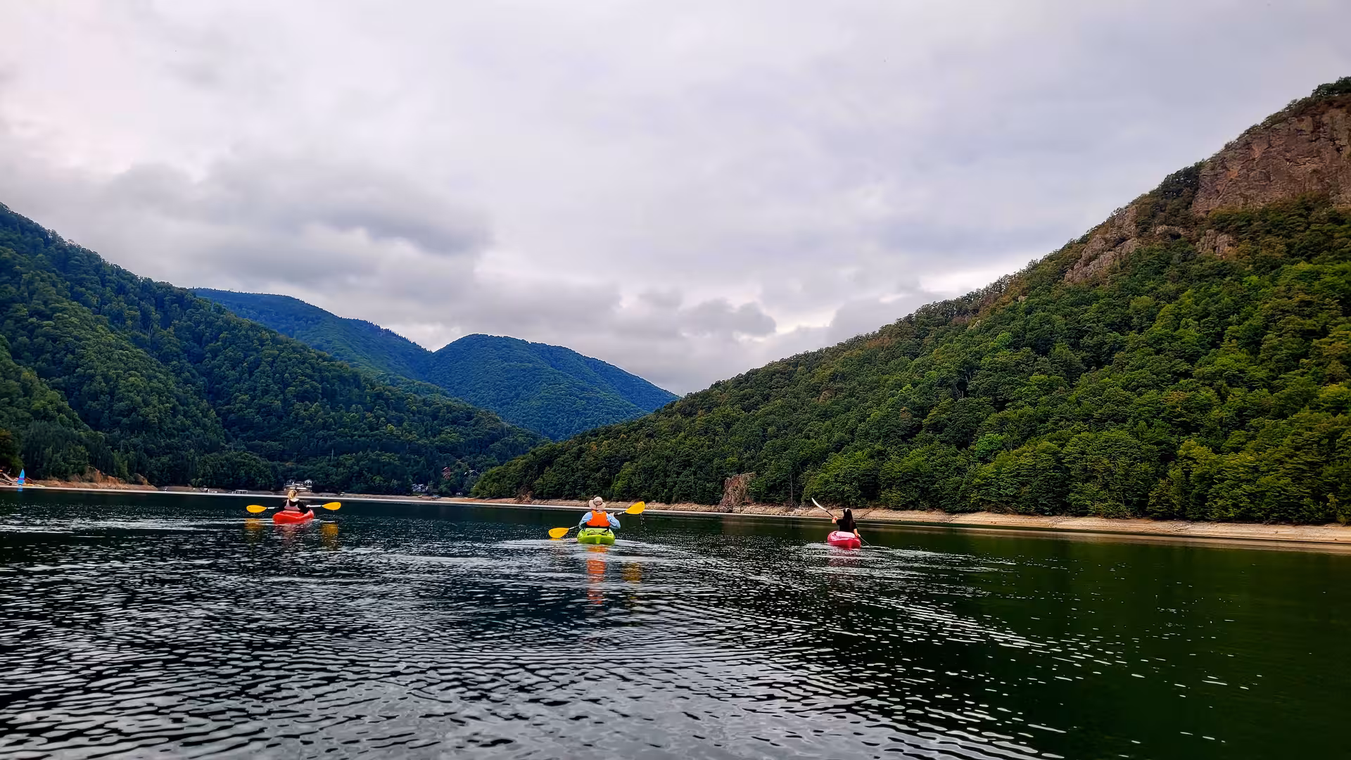 Three kayakers paddling on a calm mountain lake surrounded by lush forested Carpathian hills