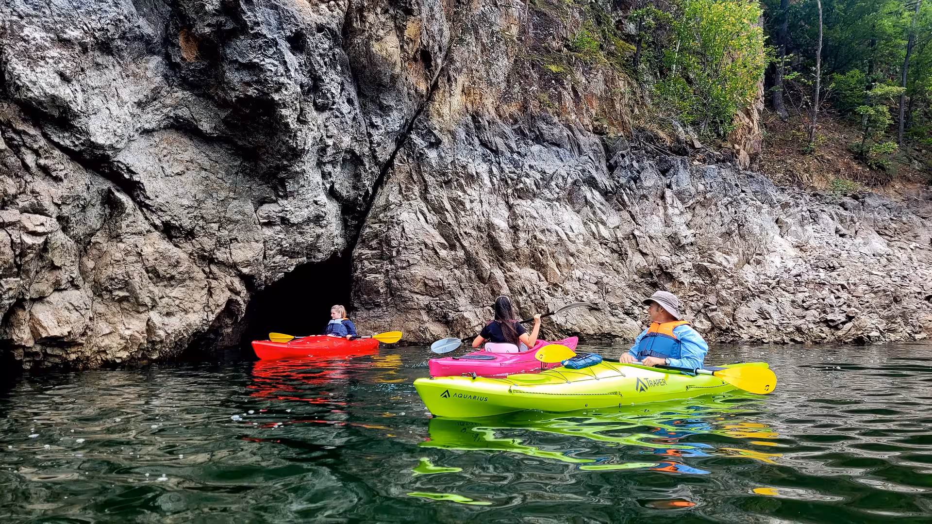 Three kayakers paddling colorful kayaks near a rocky cave entrance on a calm mountain lake