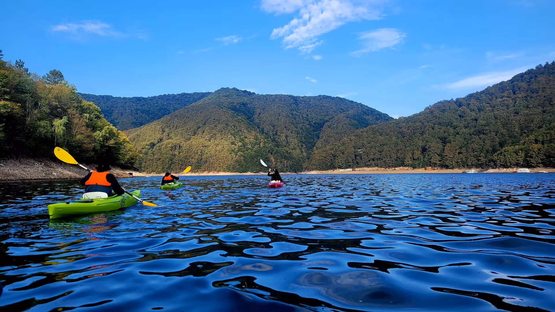Three kayakers paddling on a calm mountain lake surrounded by lush green forested hills under a blue sky