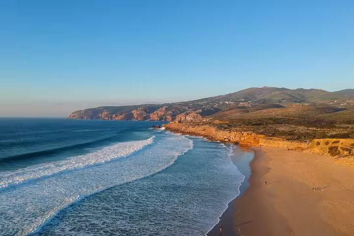 Scenic coastline of Cascais with golden sandy beach, azure ocean waves, and distant rugged cliffs under a clear blue sky.