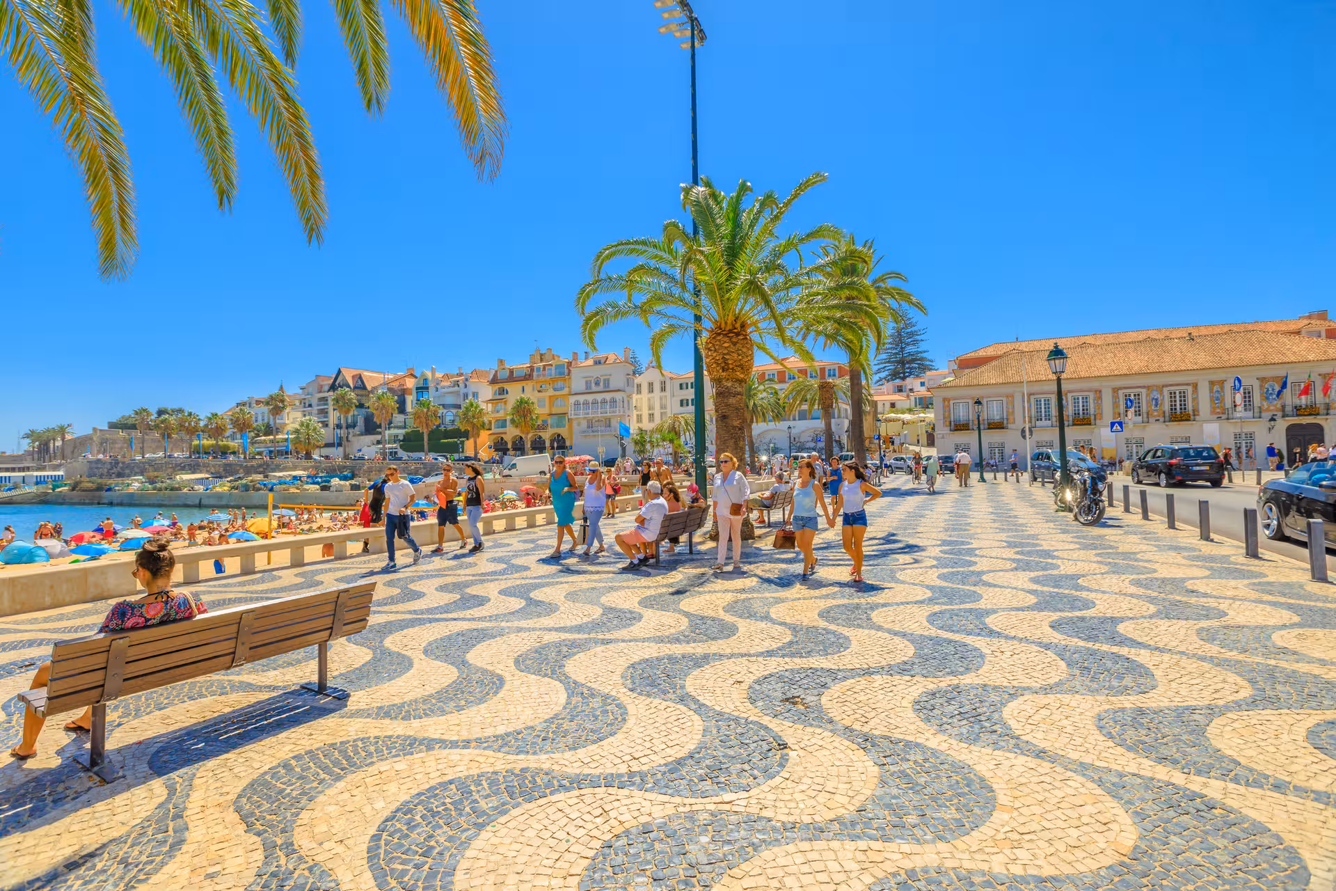 Vibrant Cascais promenade with iconic wave-patterned tiles, palm trees, and beachgoers enjoying a sunny day.