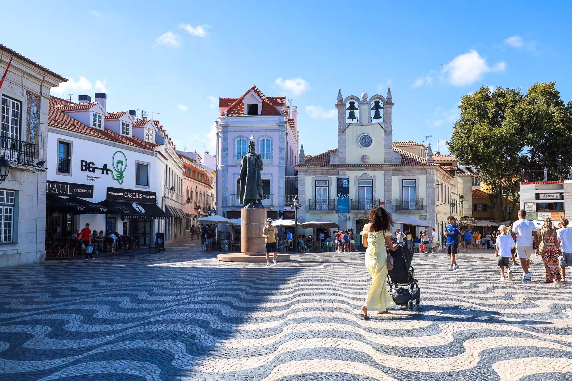 Vibrant street scene in Cascais with people enjoying cafes and a statue under a clear blue sky on a Lisbon day tour.