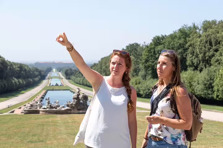 Guide leading two visitors through the Royal Palace of Caserta gardens on a sunny private tour with grand fountains behind