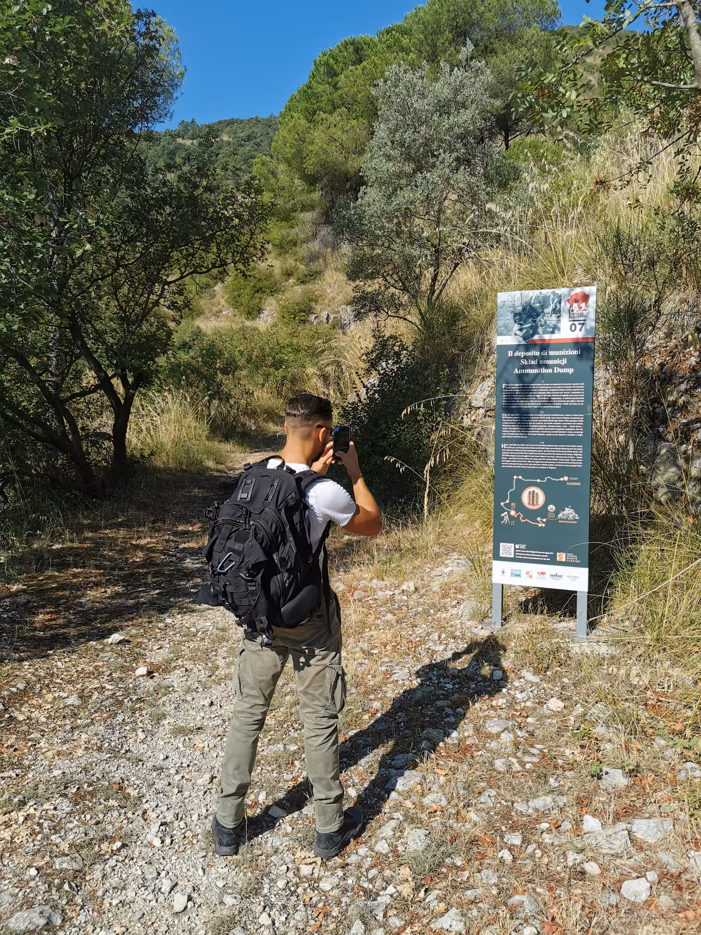 Hiker photographing an informational sign on the Cassino Red Poppy walking tour trail surrounded by lush greenery.