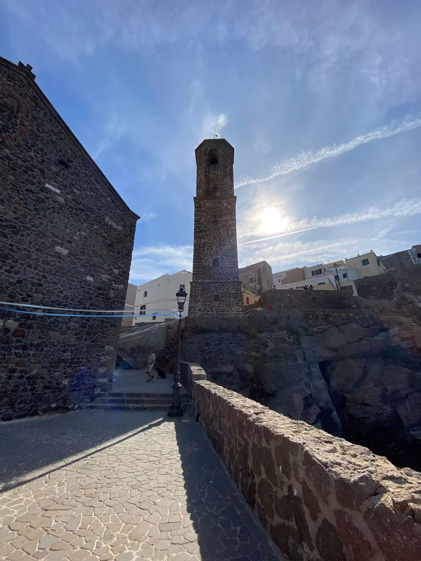 Historic tower in Castelsardo with sunlit sky, surrounded by ancient stone buildings and pathways.
