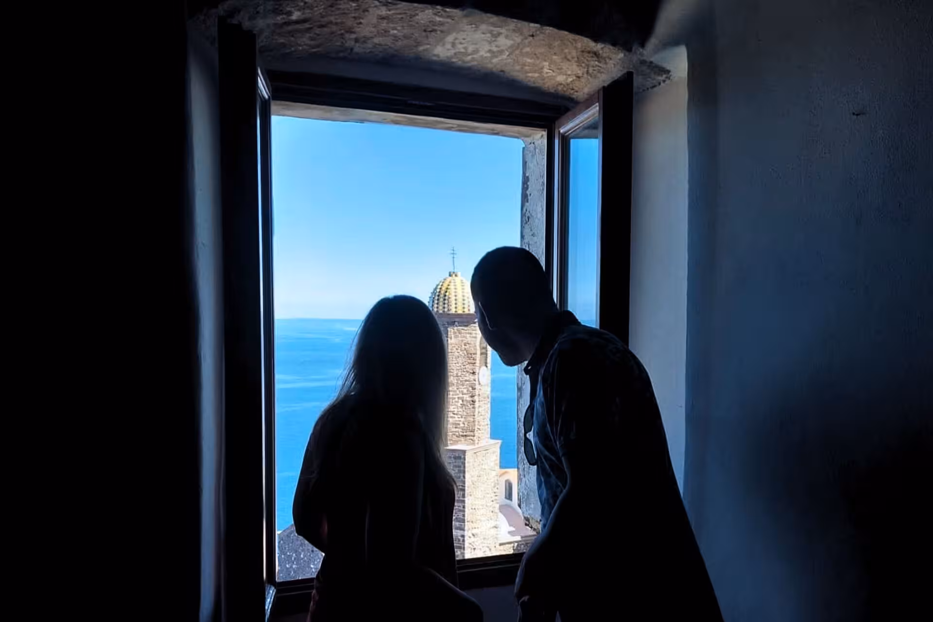 Silhouetted couple enjoying scenic view of Castelsardo's historic tower and blue sea from a window.