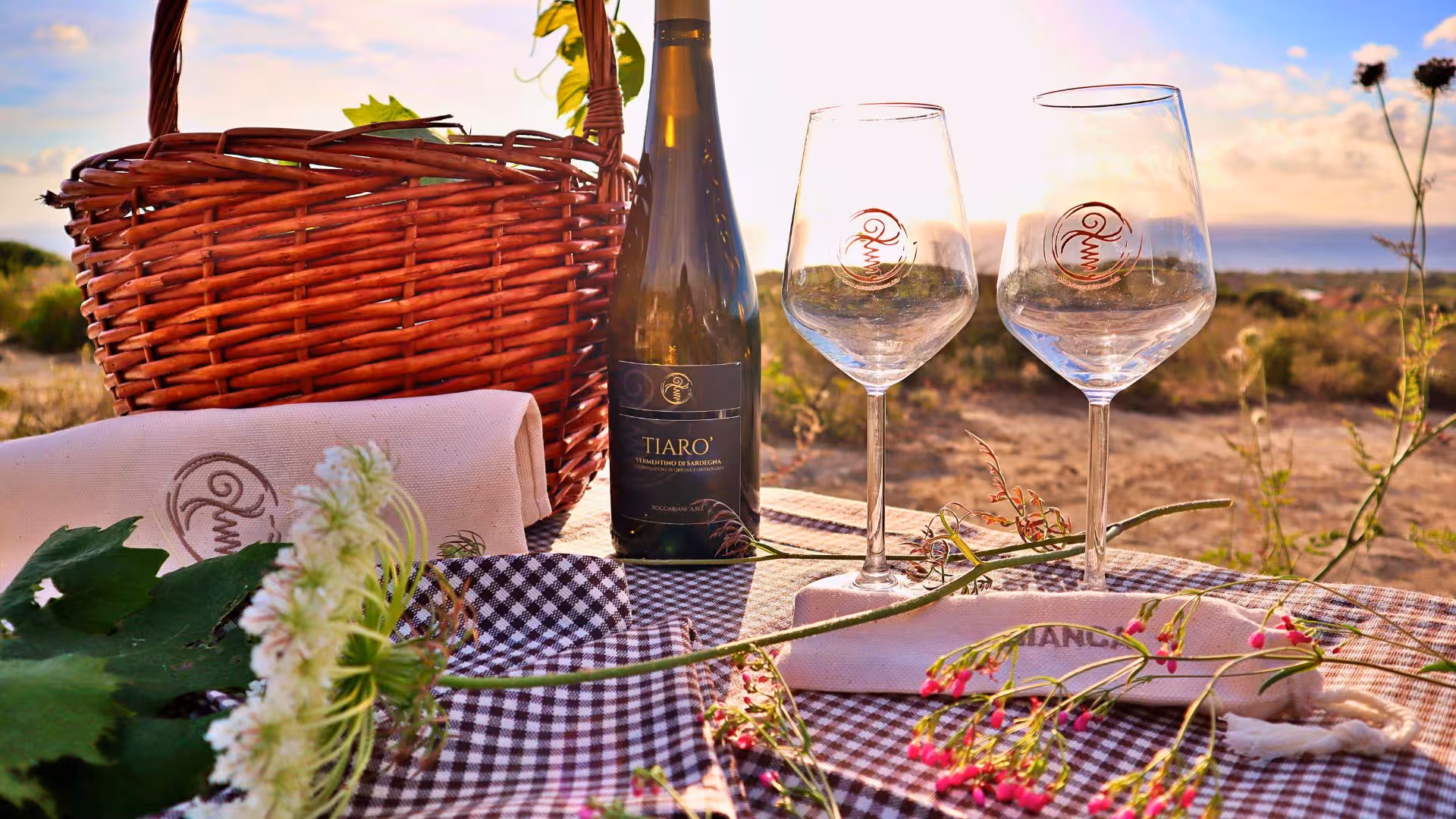 Picnic setup with wine bottle, glasses, and basket on a checkered cloth overlooking the sea in Castelsardo estate.