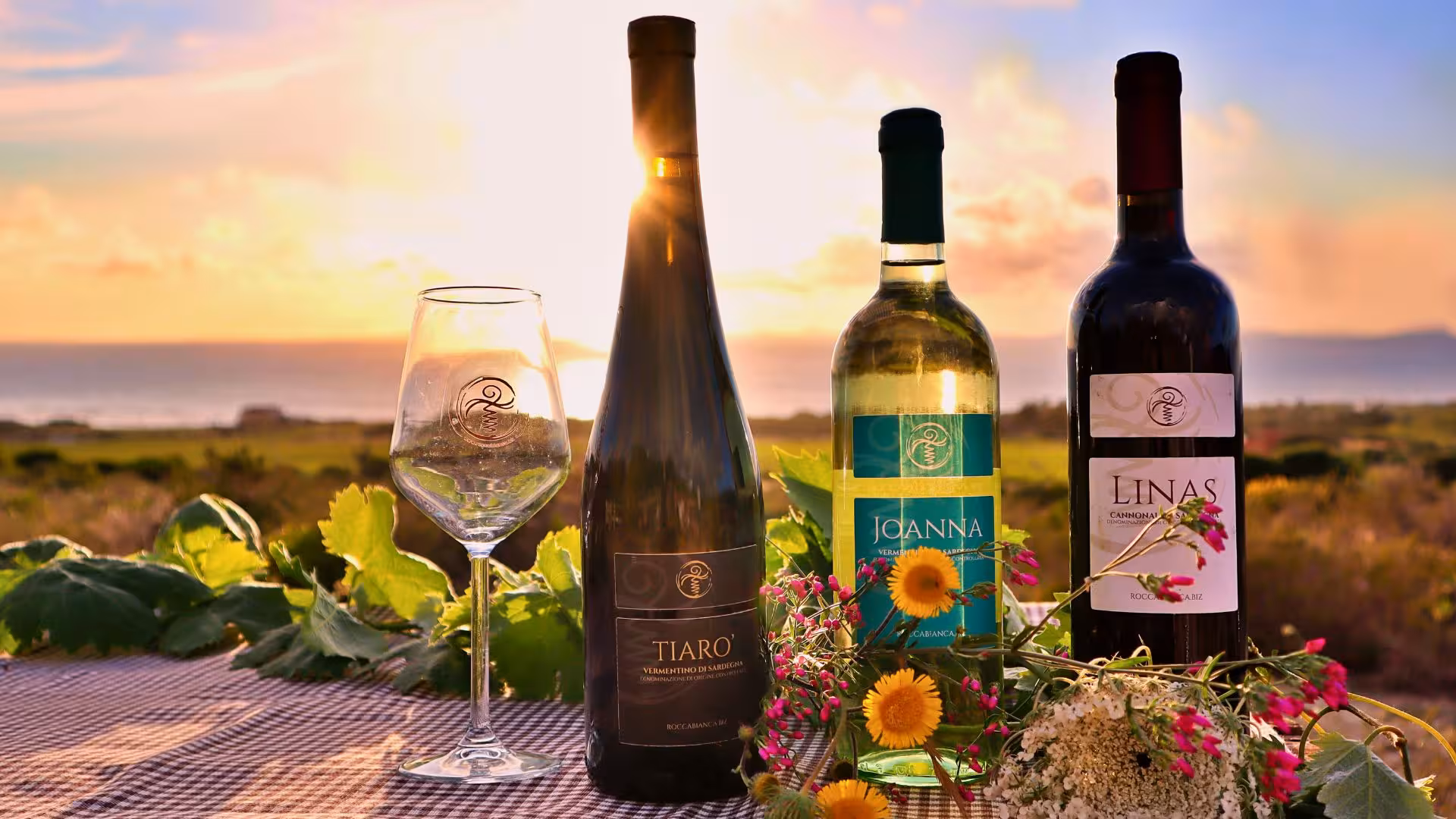 Sunset view of wine bottles and glass on a Castelsardo vineyard table with vibrant flowers and ocean backdrop.