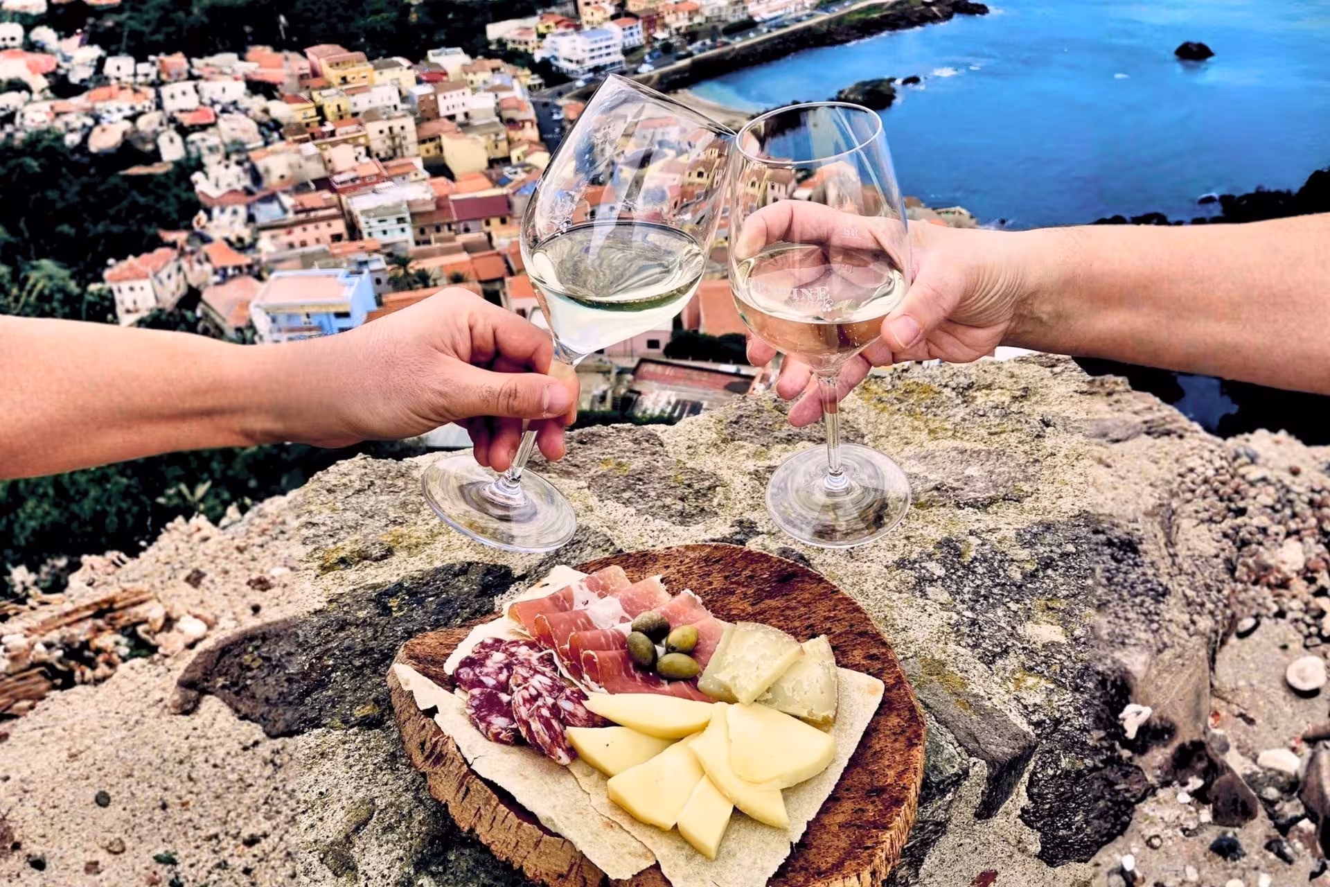 Two people clink wine glasses over a platter of local delicacies with a scenic view of Castelsardo's coastline.