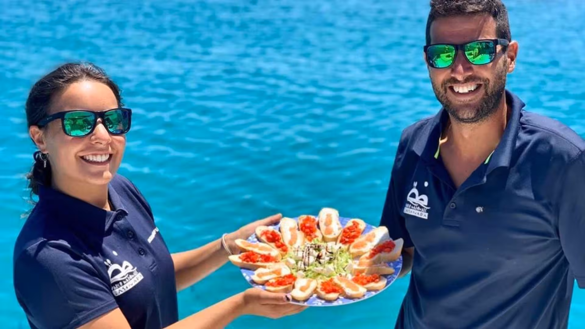 Smiling crew presenting a plate of delicious appetizers aboard a catamaran in the Gulf of Asinara, with turquoise waters in the background.