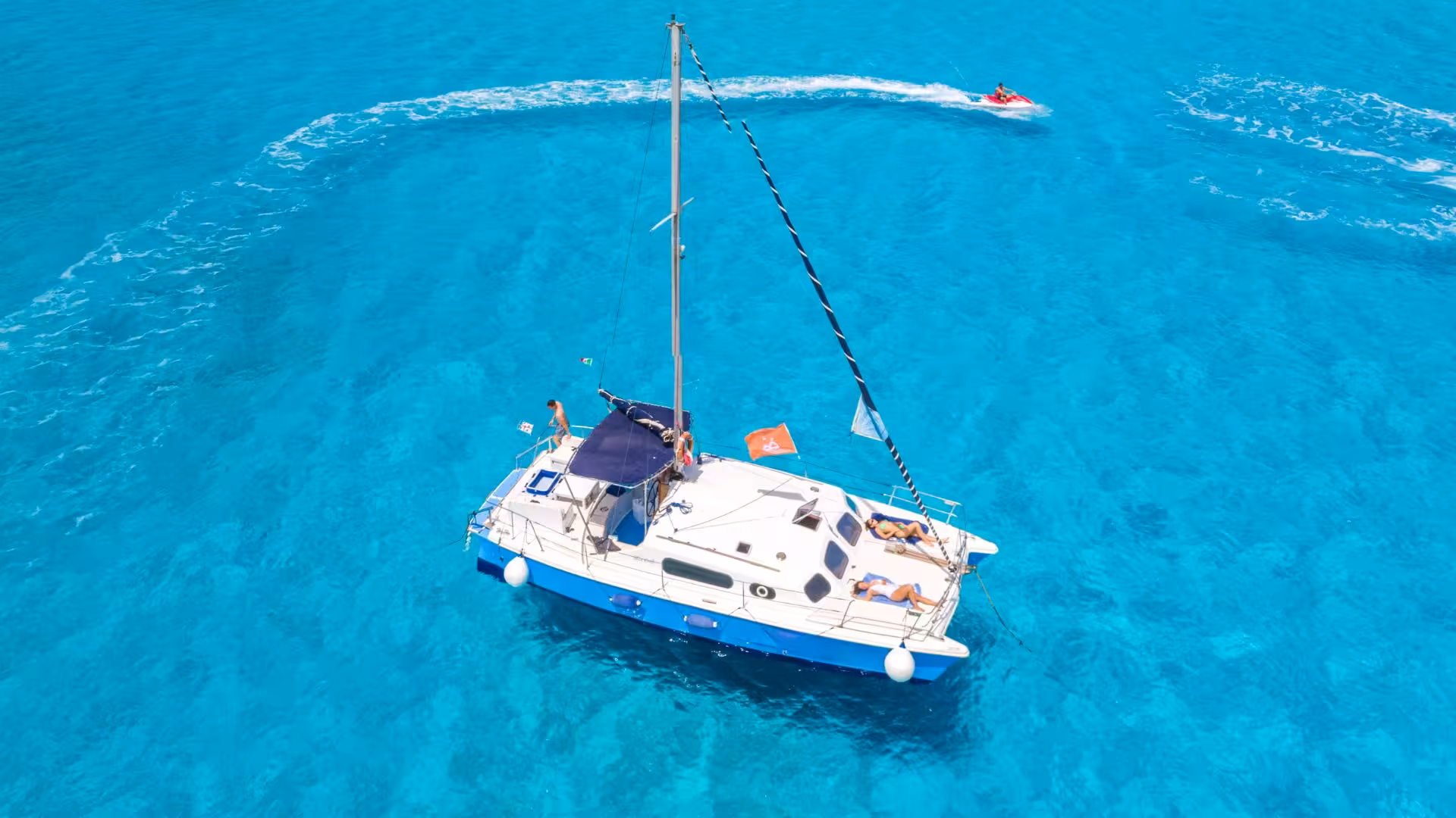 Aerial view of a catamaran sailing in the vibrant blue waters of the Gulf of Asinara, with a jet ski in the background.