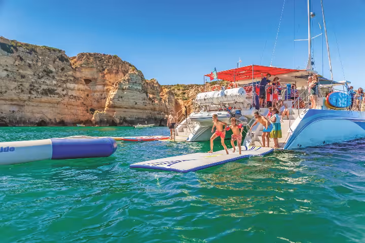 Kids jumping into crystal-clear green waters from a Lagos catamaran tour boat near stunning Algarve sea caves