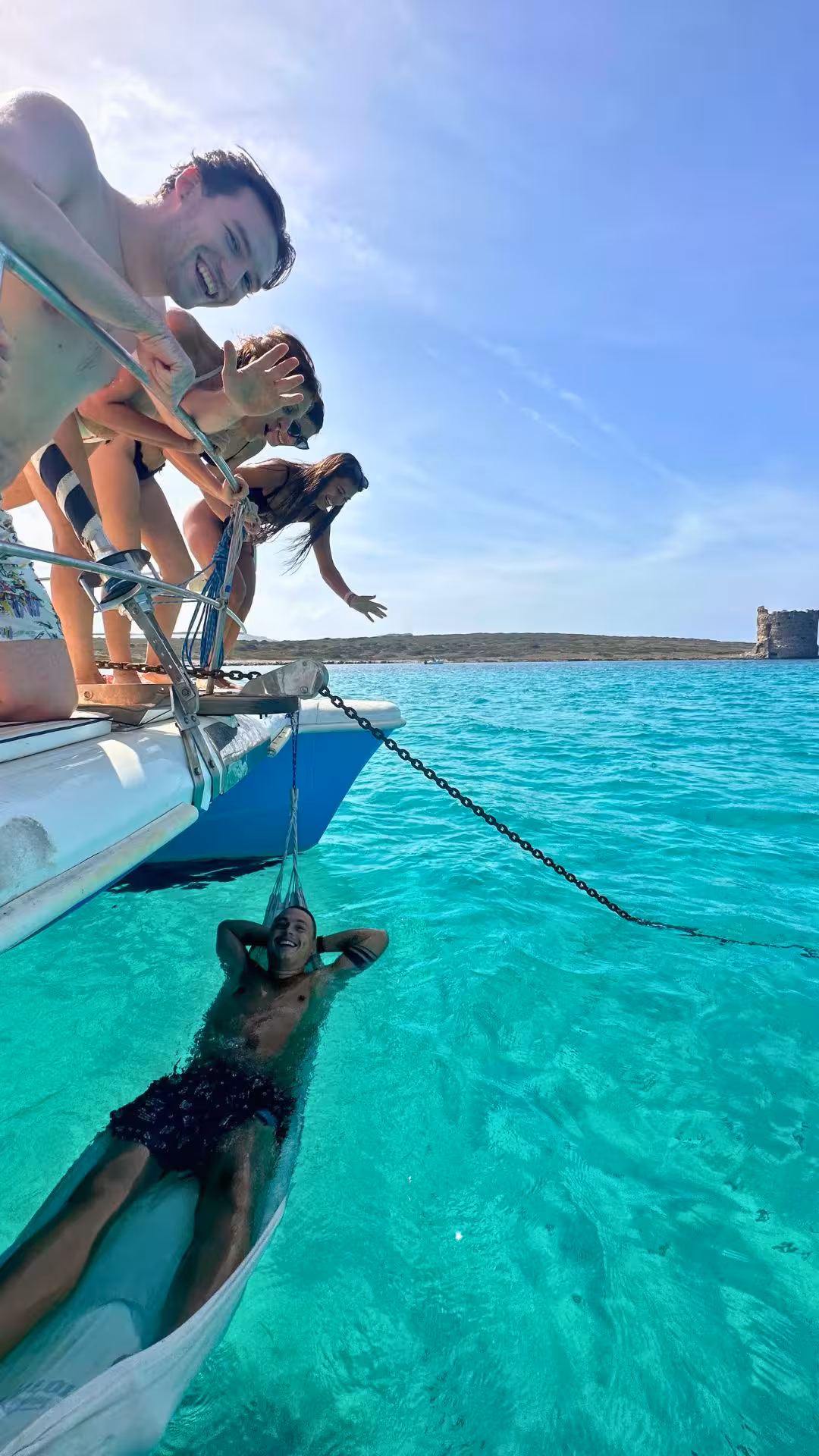 Group of friends having fun on a catamaran tour in the Gulf of Asinara, with one person relaxing in the turquoise sea.