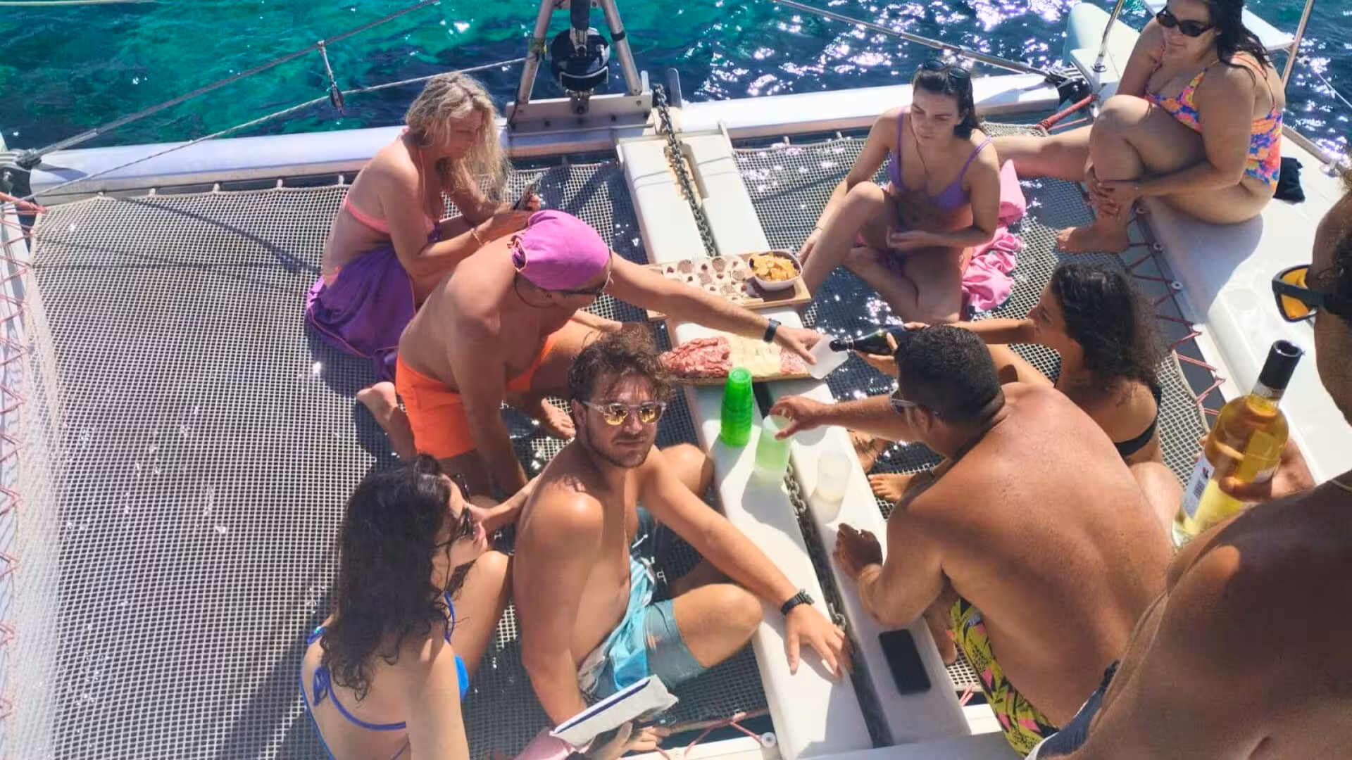 Tourists relaxing and sharing snacks on a catamaran netting, overlooking the clear blue seas of La Maddalena.