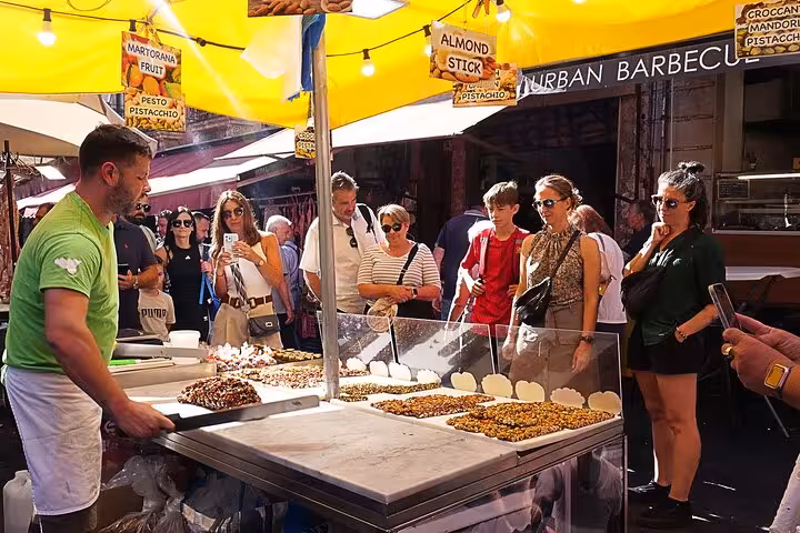 Visitors enjoy a bustling street food market in Catania, savoring a variety of Sicilian treats like almond sticks.