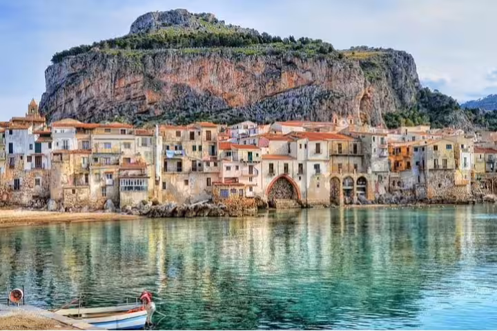 Colorful waterfront houses of Cefalù reflected in clear turquoise sea beneath dramatic rocky cliffs on a private day trip from Palermo