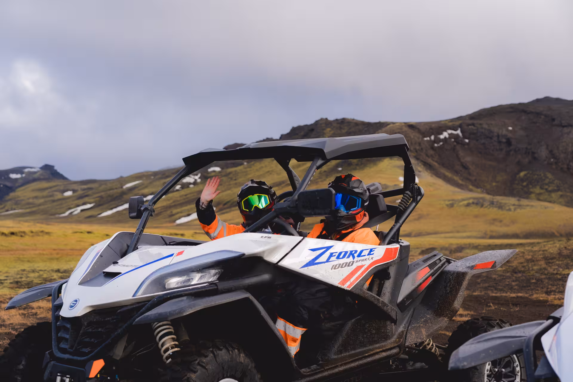 Two riders in orange gear and helmets waving from a CFMoto ZForce 1000 buggy in Iceland's green highlands