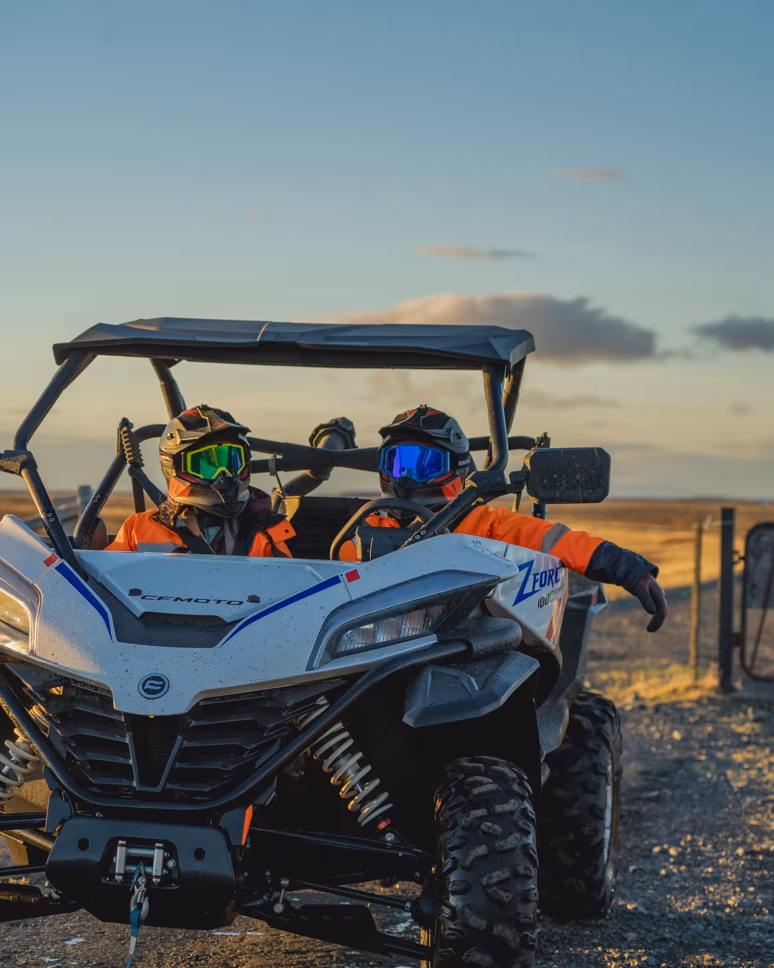 Two riders in orange gear and helmets sitting in a white CFMOTO ZForce buggy on Icelandic terrain at sunset