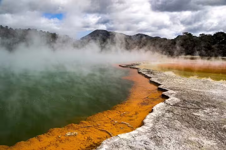 Steaming Champagne Pool in Rotorua with vivid orange and green waters against a backdrop of mountains and clouds.