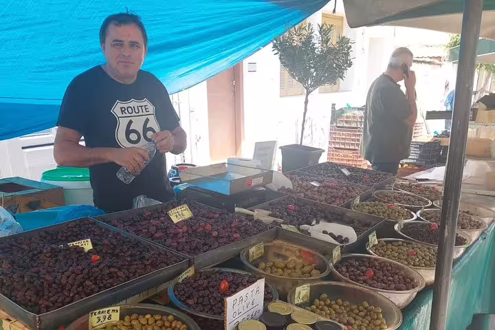 Vendor showcasing a variety of olives at Chania's Laiki market, with bowls of olives under a vibrant blue canopy.