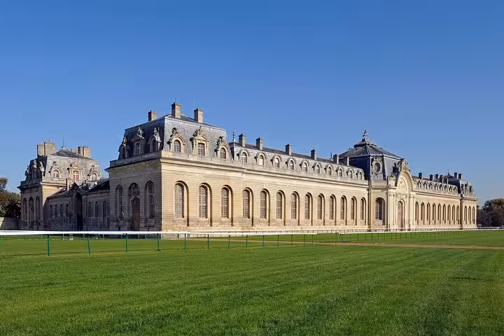 Grand façade of Château de Chantilly with manicured green lawns under a clear blue sky, France