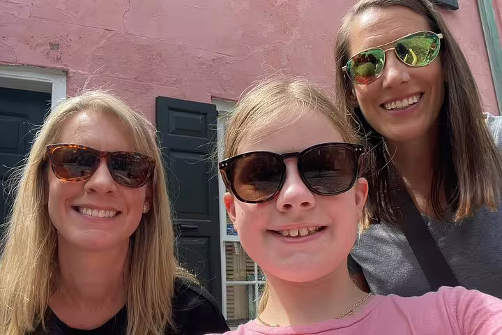 Two women and a girl wearing sunglasses smiling in front of a pink historic Charleston building