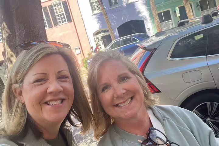 Two smiling women taking a selfie on a sunny Charleston street with colorful historic buildings