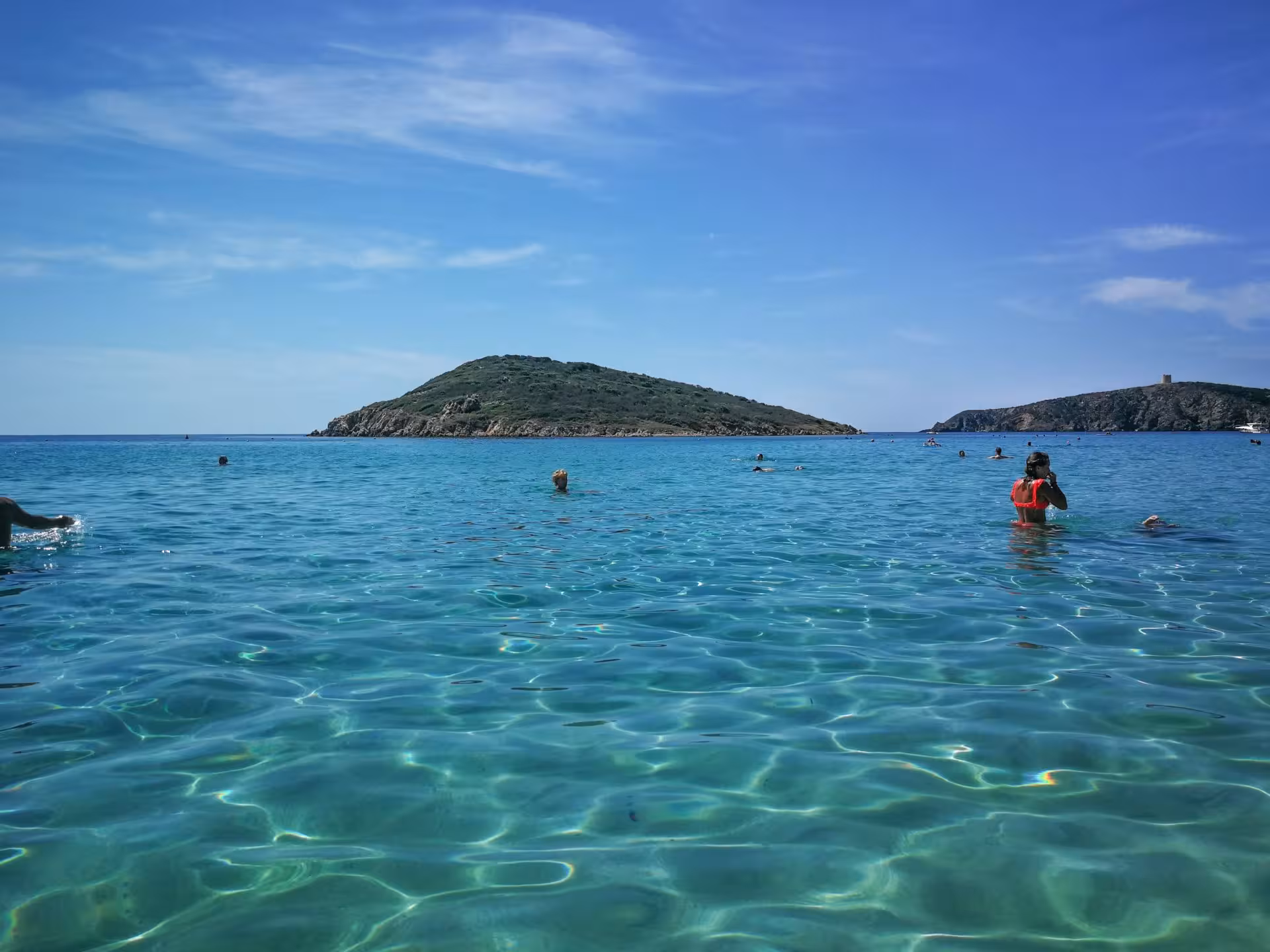 Tourists enjoy swimming in the crystal-clear waters of Chia beach, Sardinia, under a vibrant blue sky.