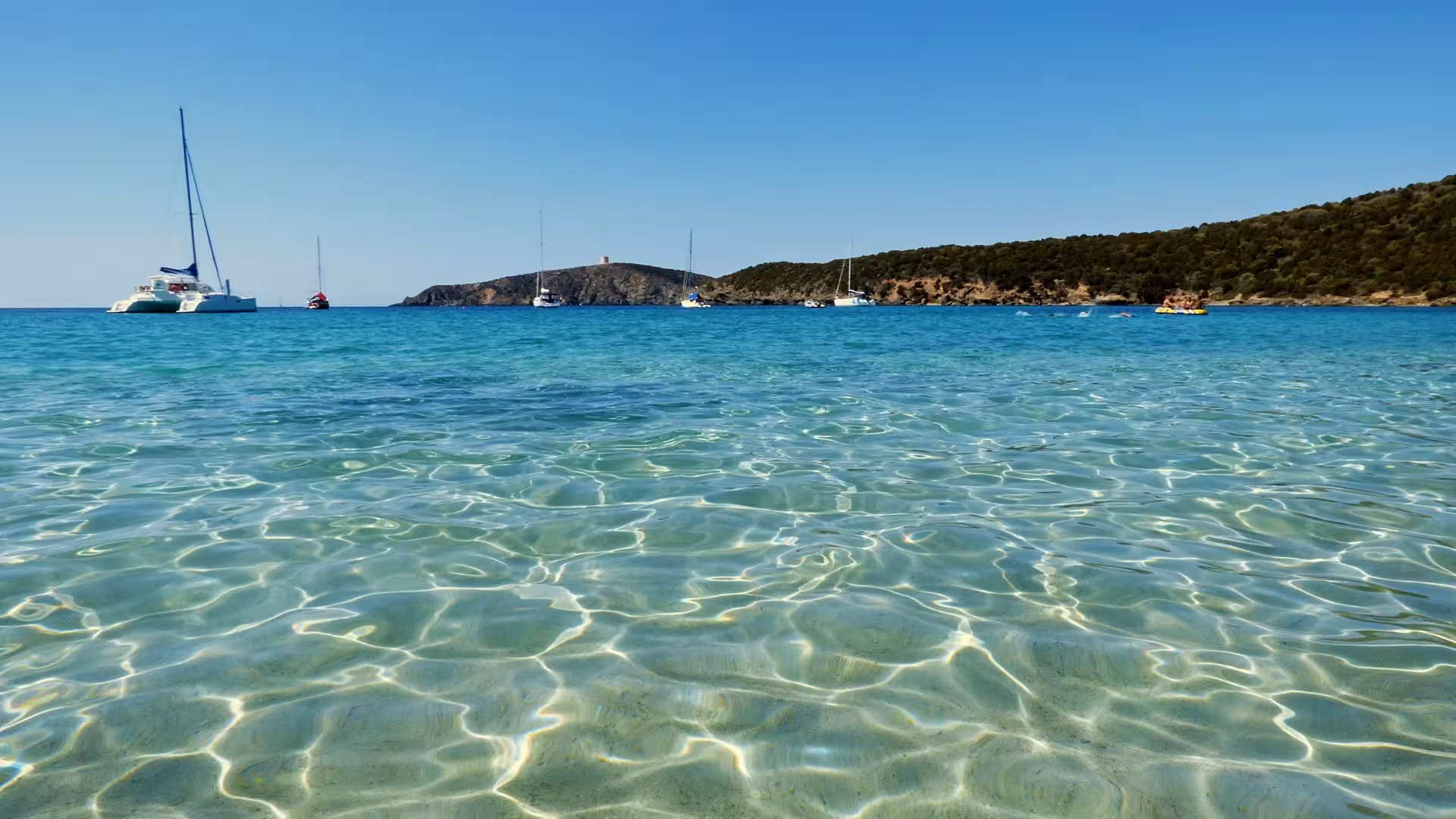 Crystal-clear waters with yachts in the distance near Chia, offering a serene setting for a dinghy tour adventure.