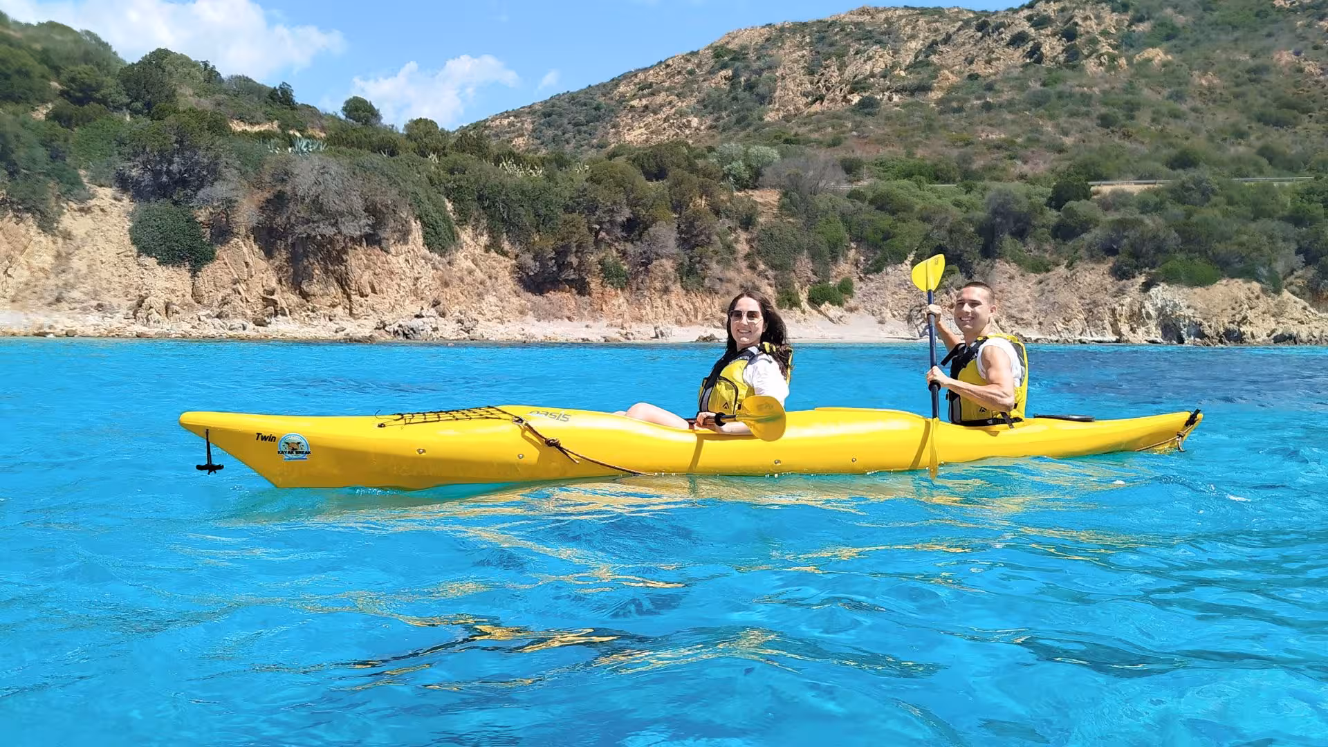 Two people kayaking on turquoise waters with rocky cliffs in the background at Tuerredda beach.