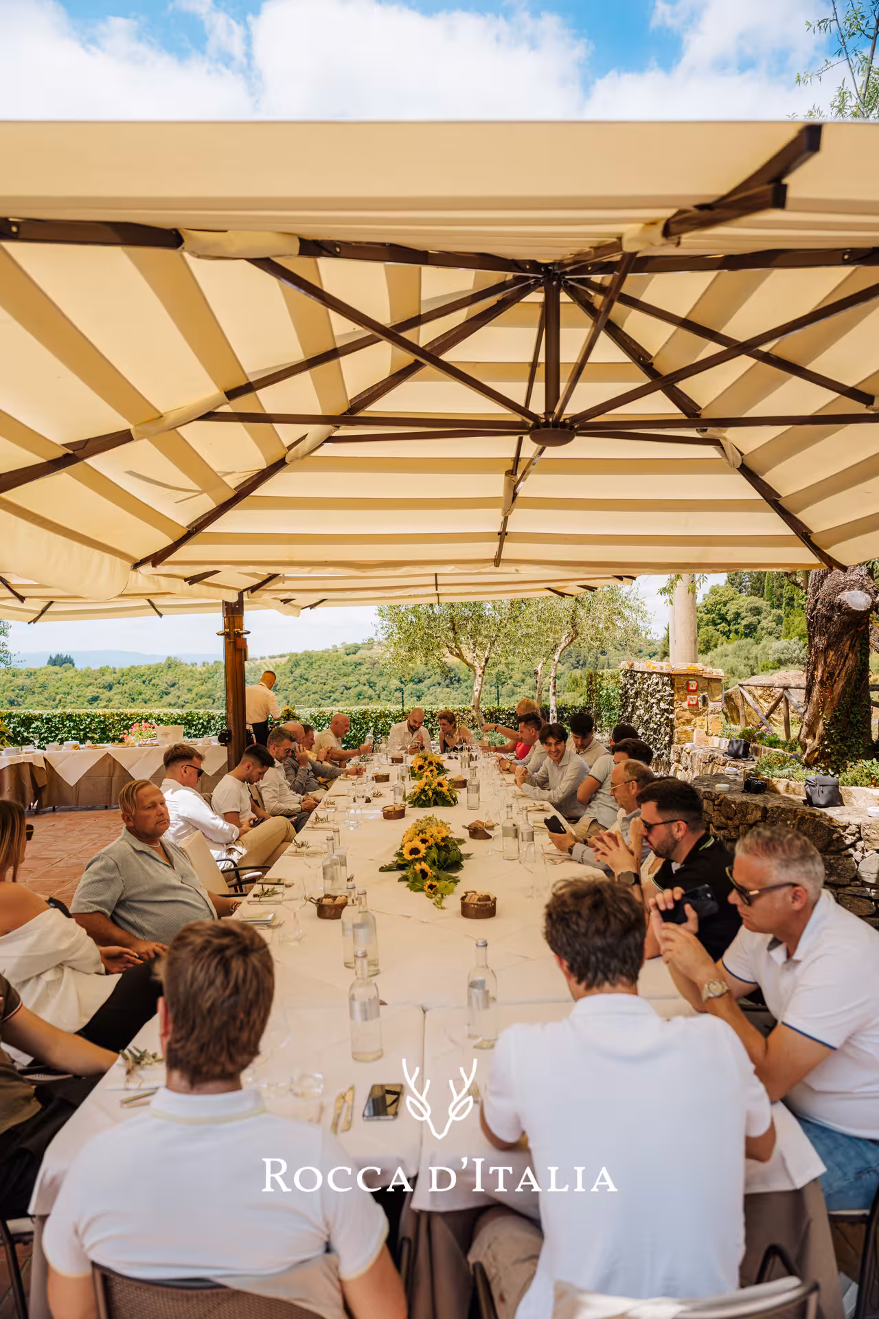Group dining outdoors at long table with sunflowers under large umbrella at Chianti vineyard terrace