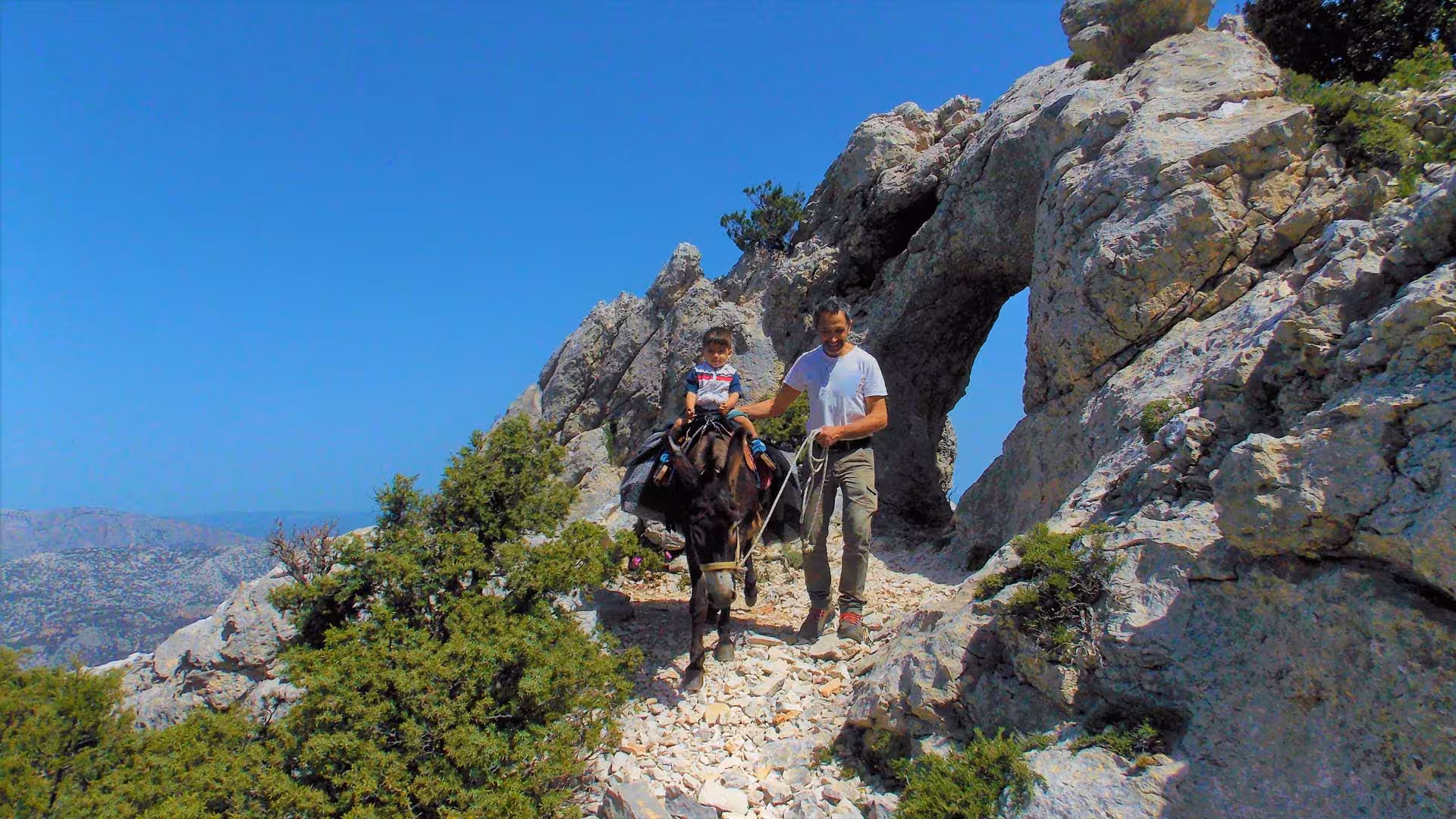 Child riding a donkey led by a guide on a rocky path with stunning Sardinian landscapes in Dorgali.