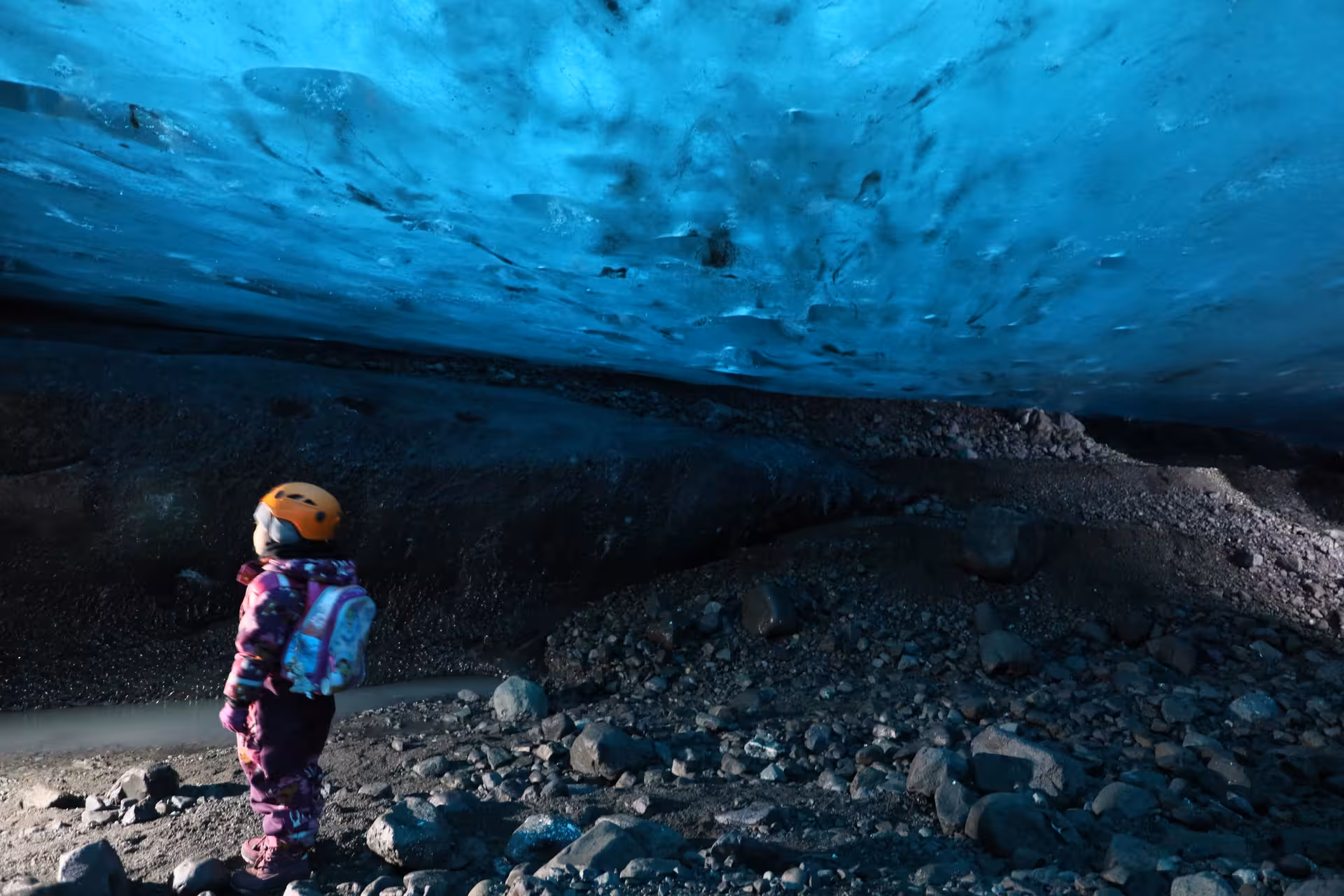 Child in orange helmet gazing up at vivid blue glacial ice cave ceiling in Iceland