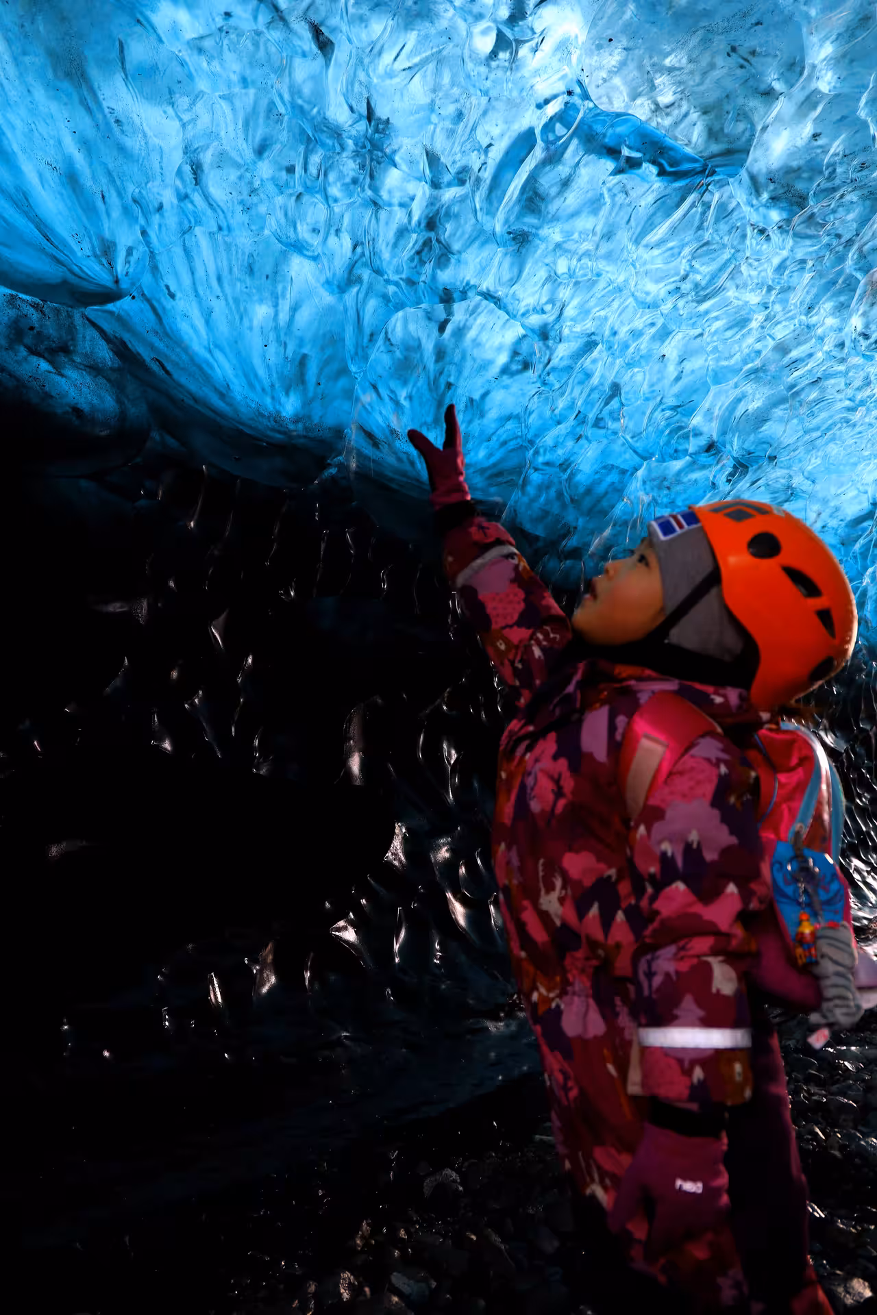 Child in orange helmet and pink suit touching blue ice cave ceiling during Iceland glacier tour