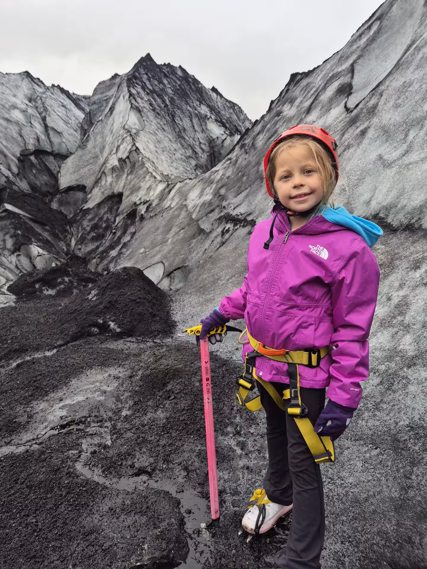 Young girl in safety harness and helmet holding pink ice axe on Iceland glacier