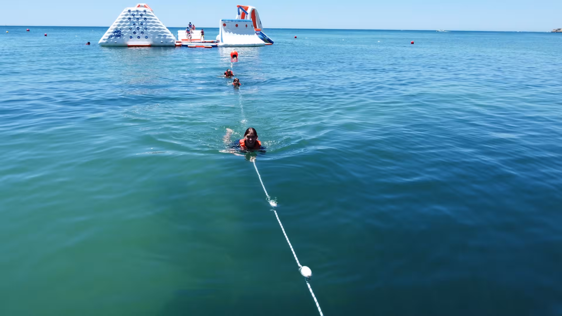 Child in life jacket swimming toward inflatable waterpark obstacles floating on calm ocean on a sunny day