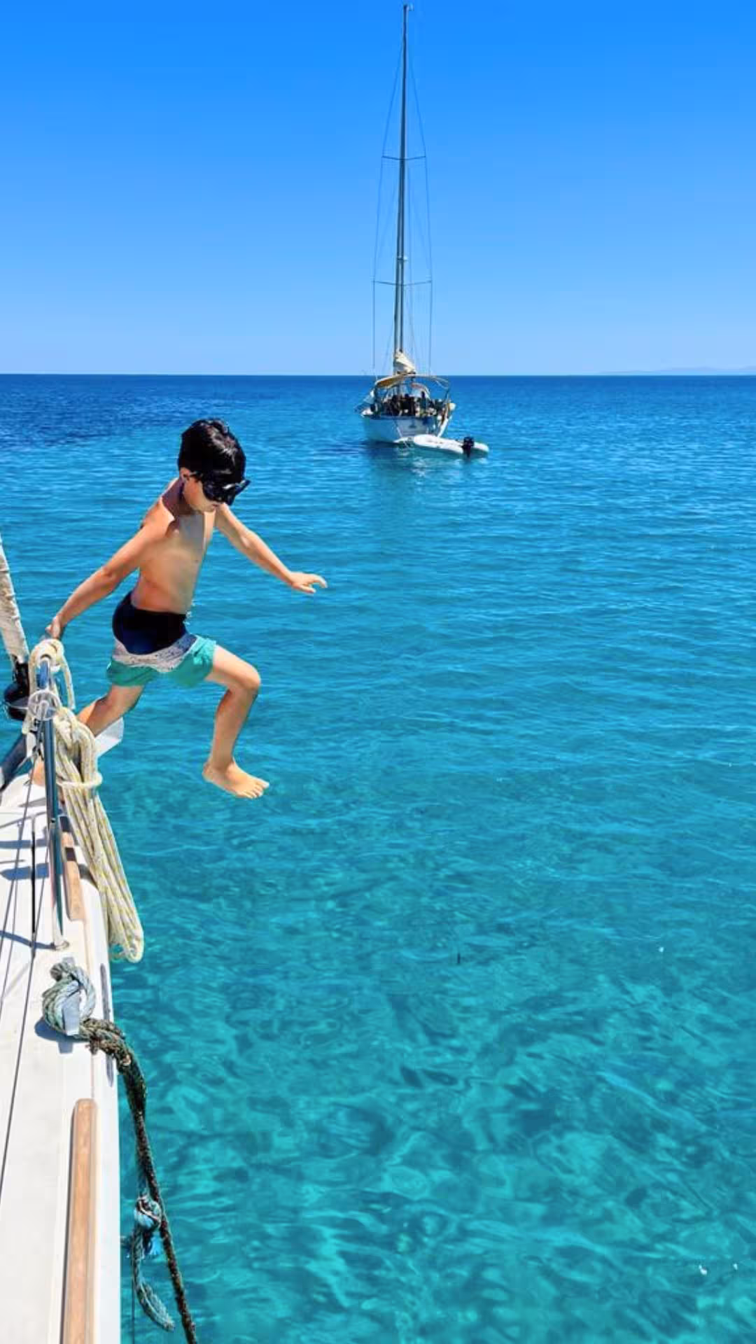 Child jumping into crystal-clear sea from a boat on an Alghero sailing tour, with a sailboat in the background.