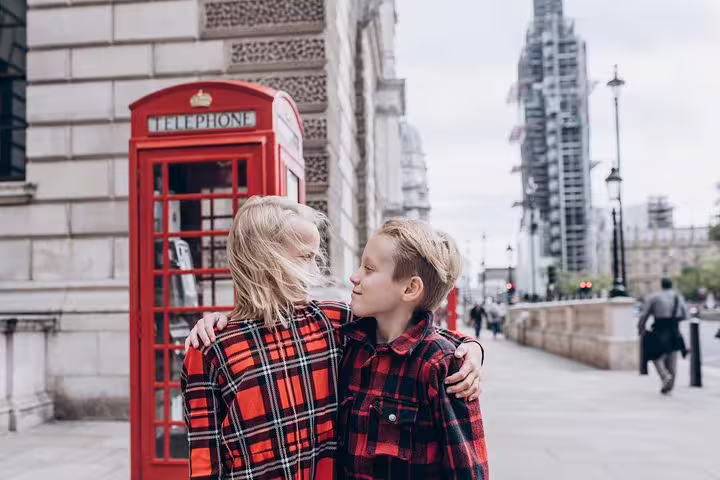 Two children in matching plaid shirts embrace near a classic red London phone booth with Big Ben in the background.