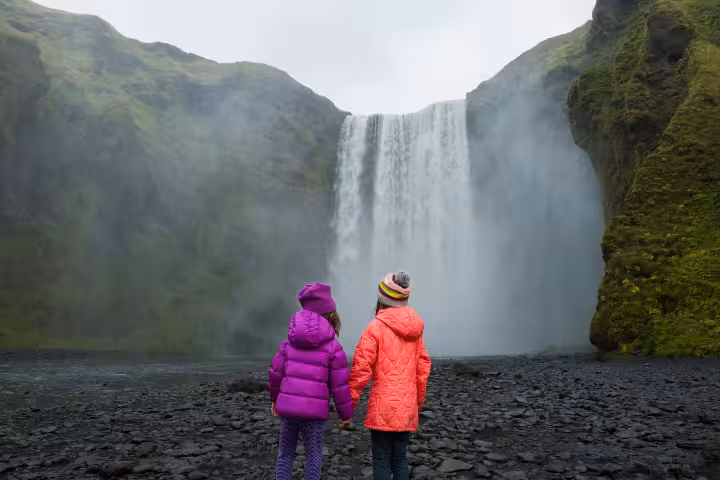 Two children holding hands facing Skógafoss waterfall on Iceland's south coast