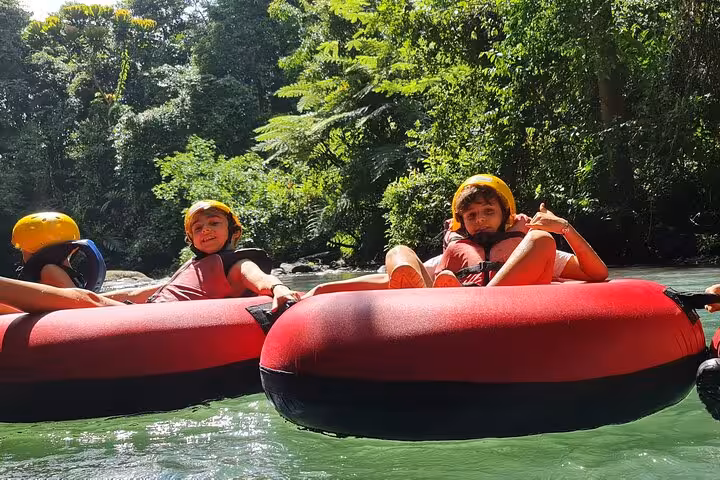 Two children wearing helmets and life jackets tubing on Rio Celeste, surrounded by lush tropical forest.