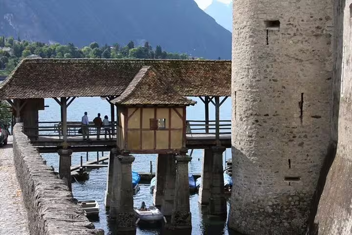 Medieval covered bridge on stilts over lake at Chillon Castle with visitors and mountains behind